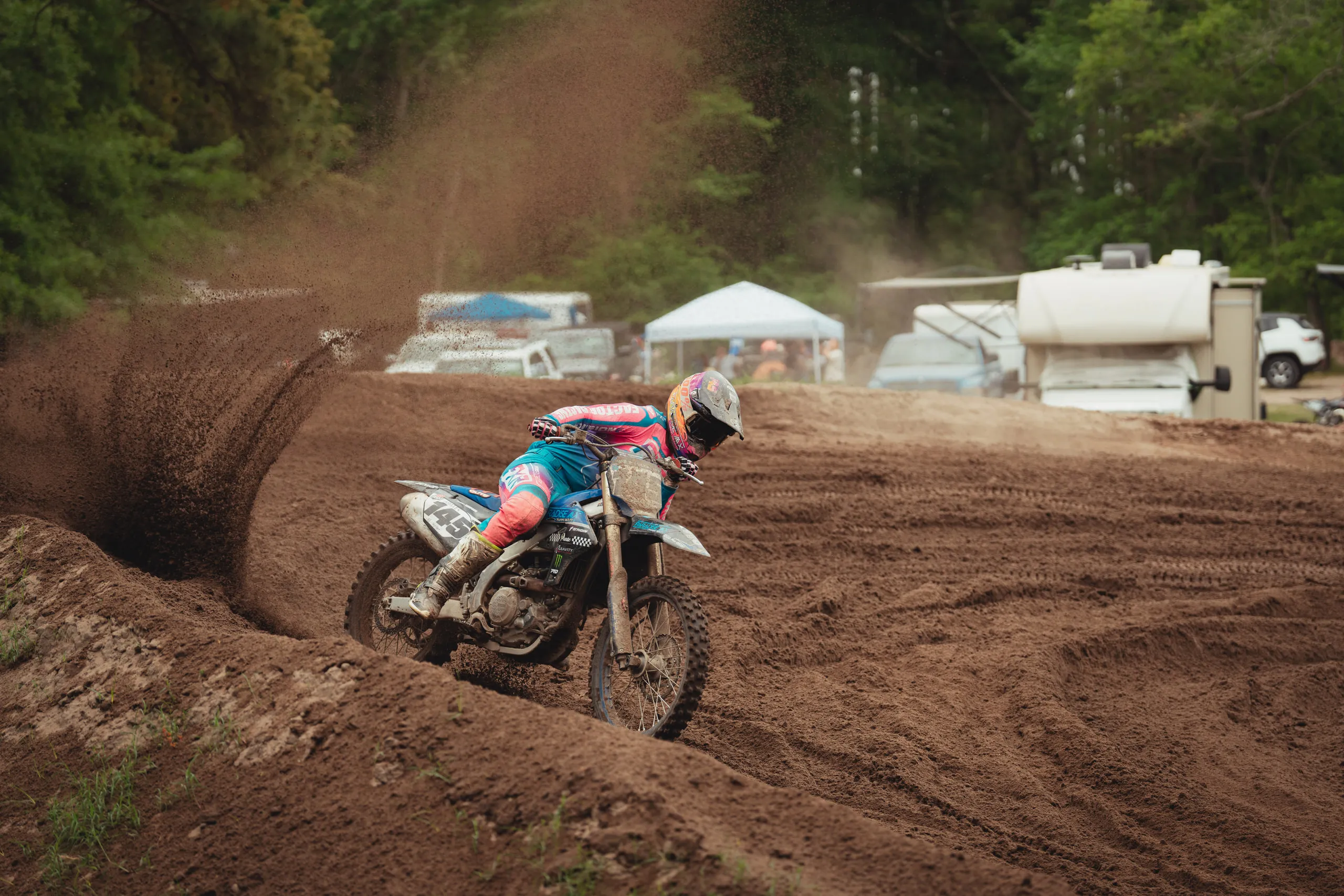 Rider leans hard through a deep berm, throwing dirt, with pit tents in the background