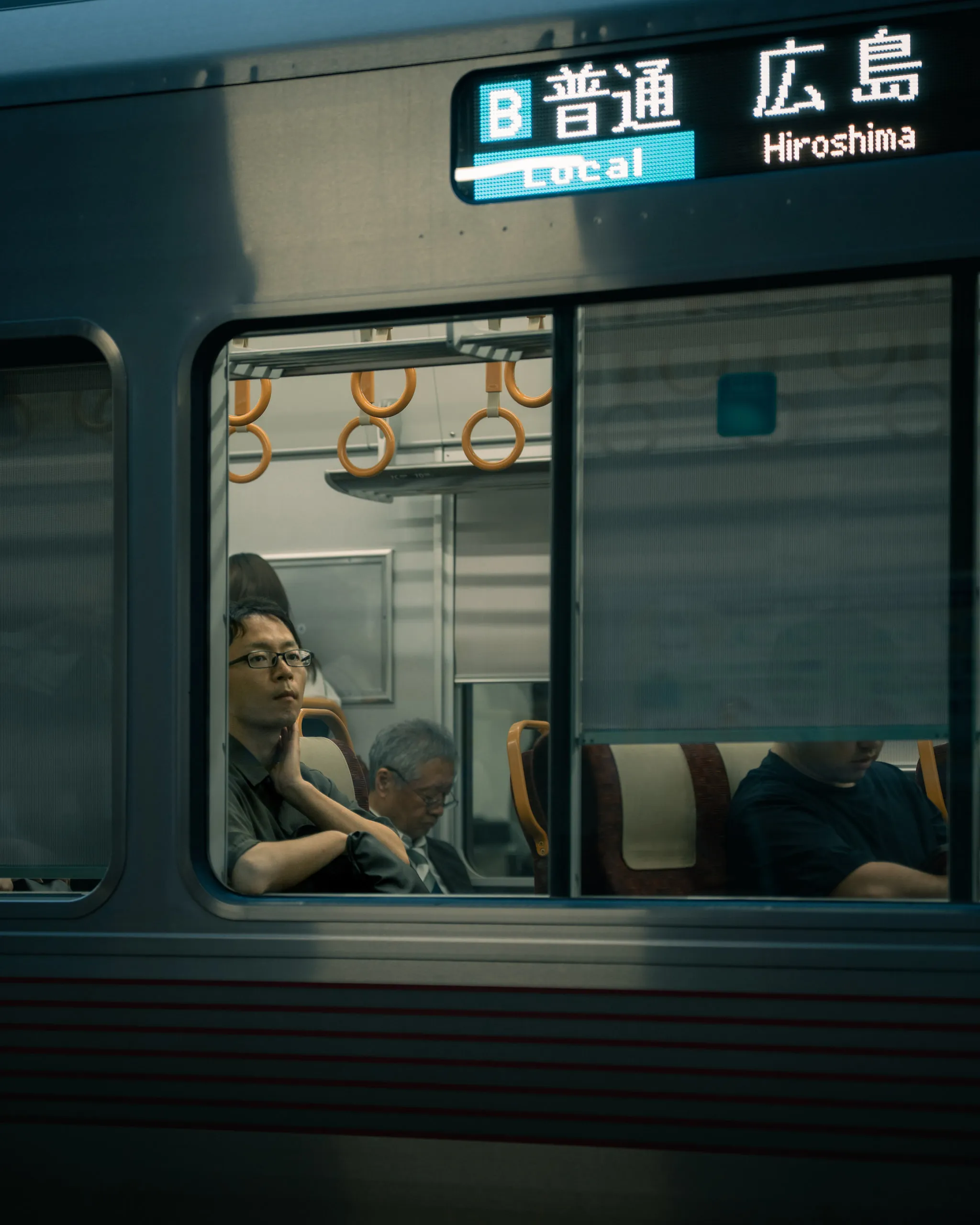 A passenger rests against the window of a local Hiroshima train, seen from the platform