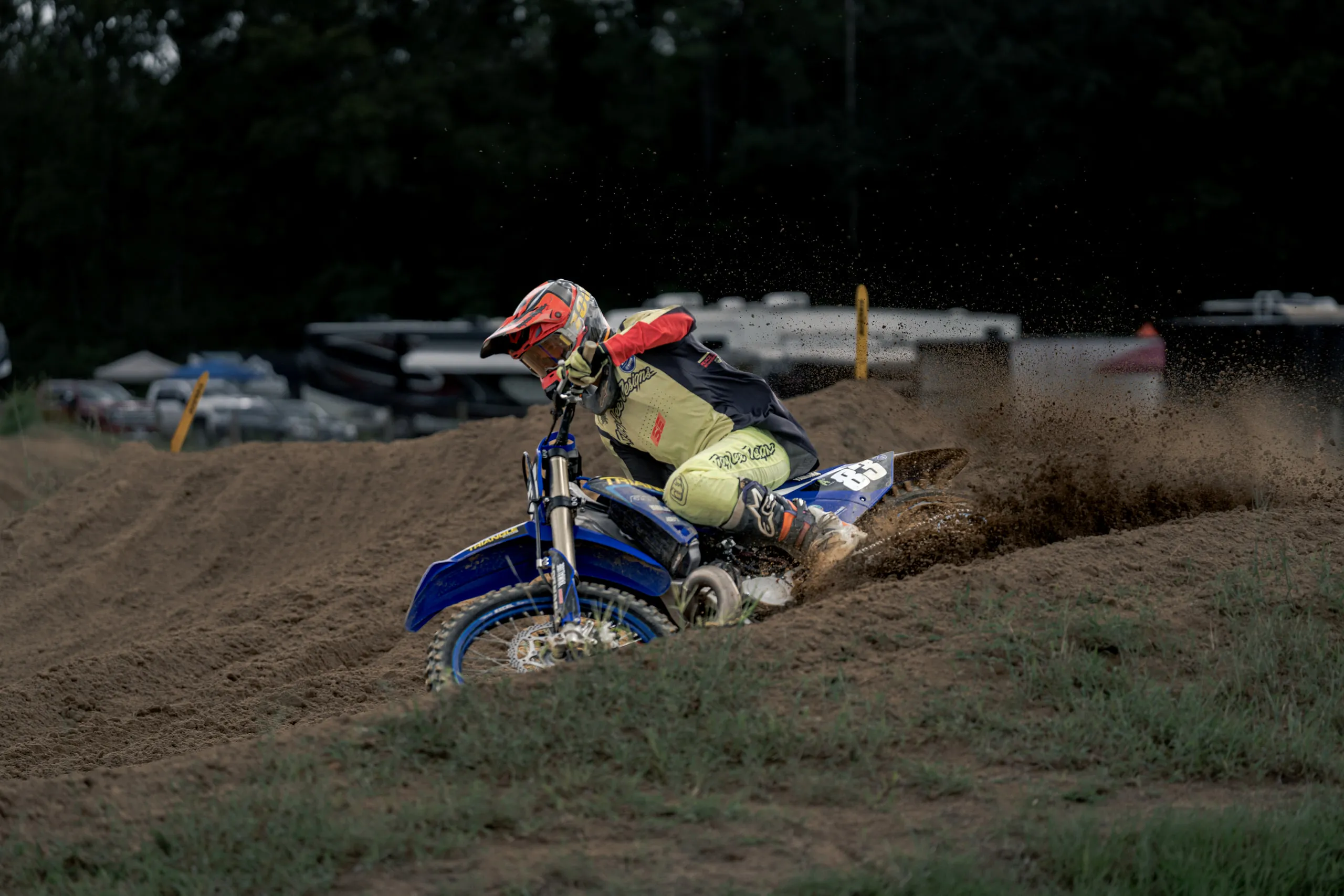 A rider in yellow gear drives a blue Yamaha through a rutted corner, throwing a sheet of dirt off the rear wheel