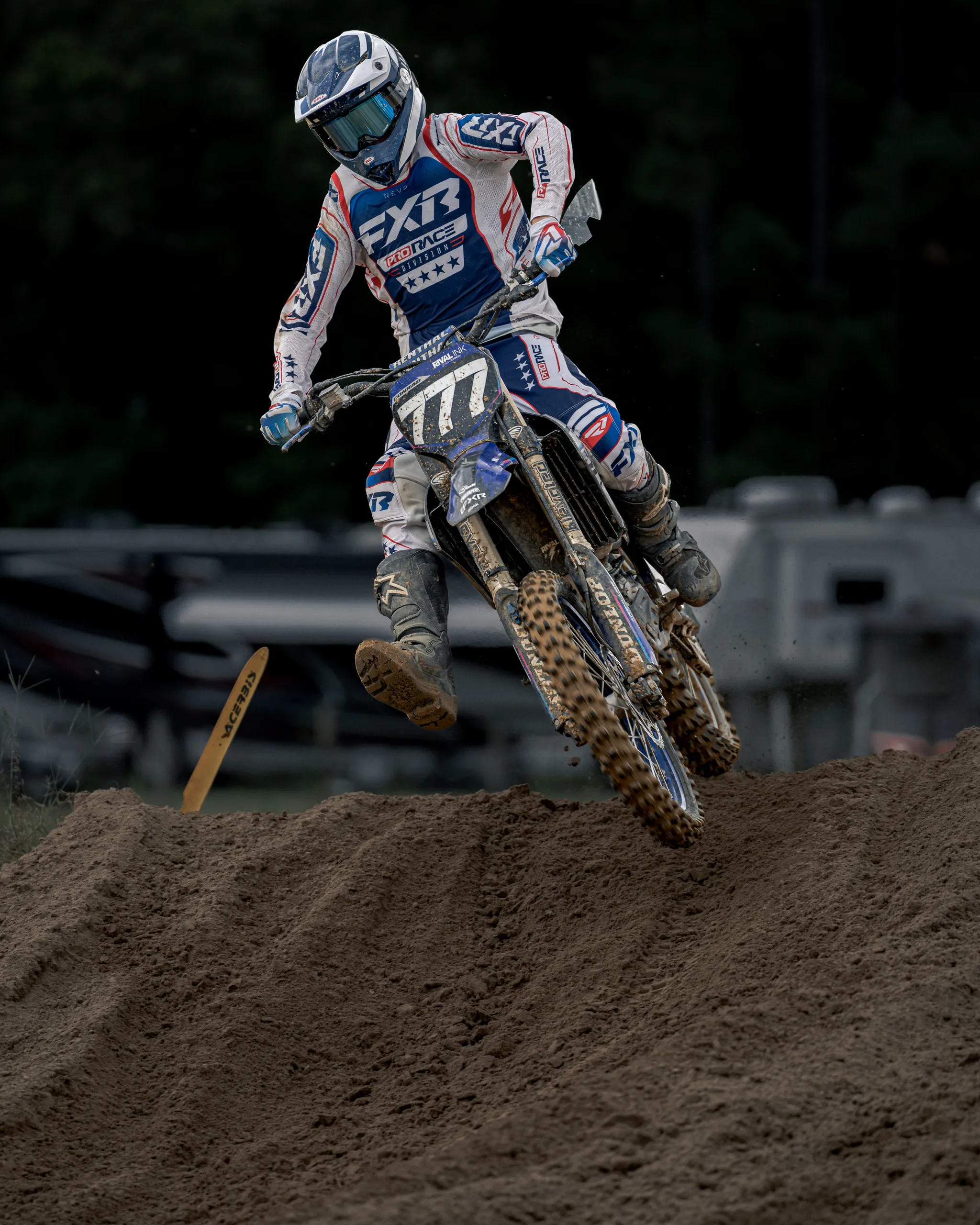 A rider in white and blue gear lifts off the face of a jump, legs braced, with pit trailers lined up behind the track