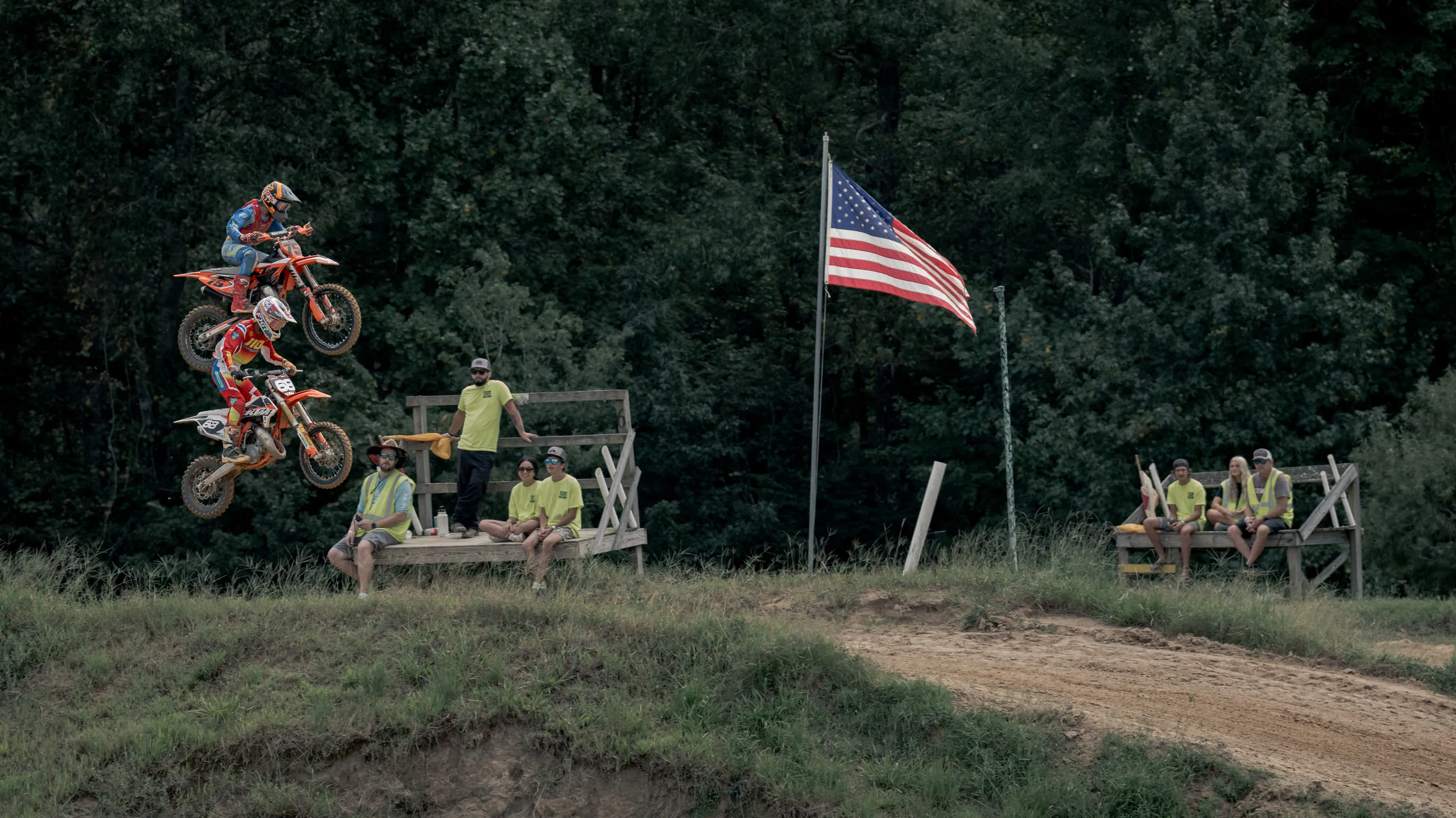 A rider sails over a jump past an American flag while spectators in hi-vis shirts watch from a wooden stand