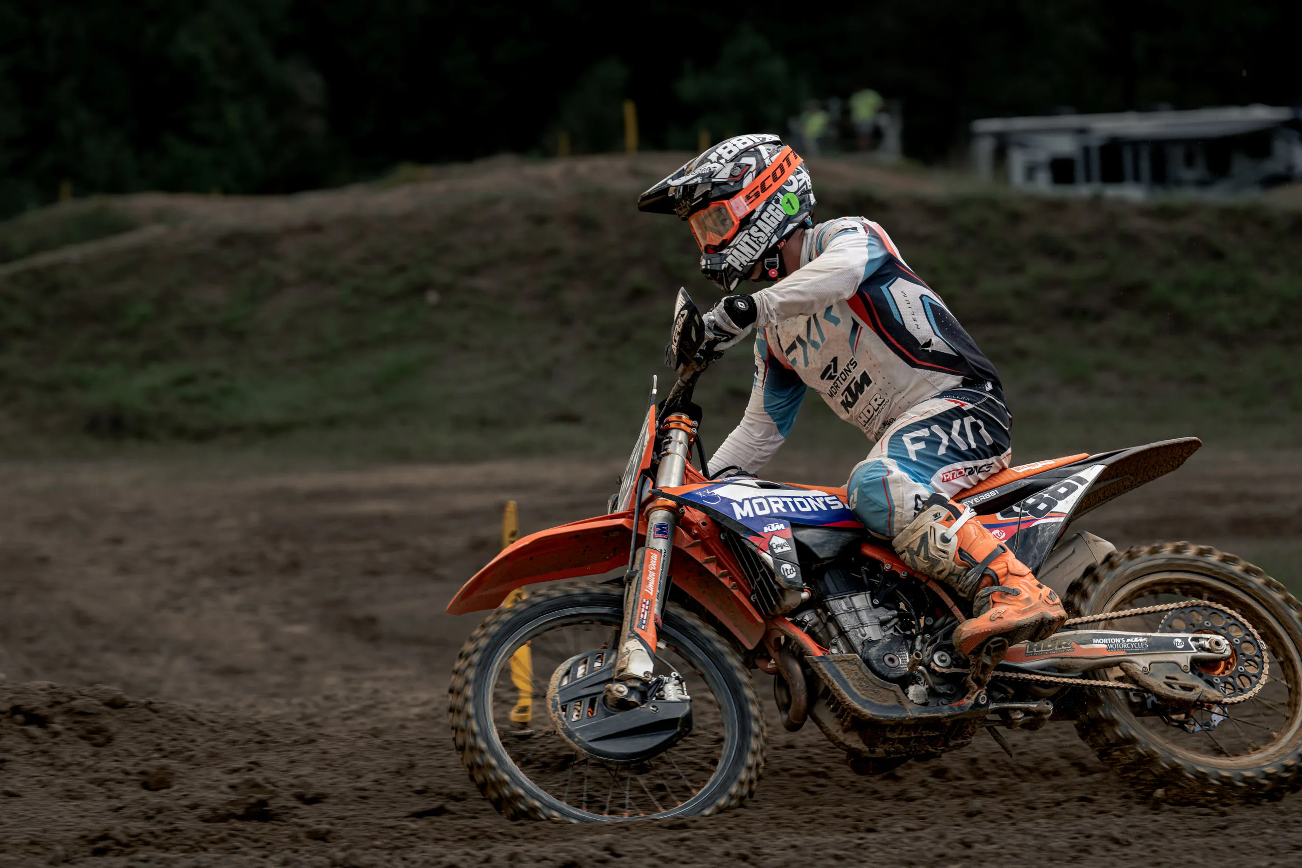 A rider leans an orange KTM through a bermed corner, shot low from trackside with the pits parked on the bank above