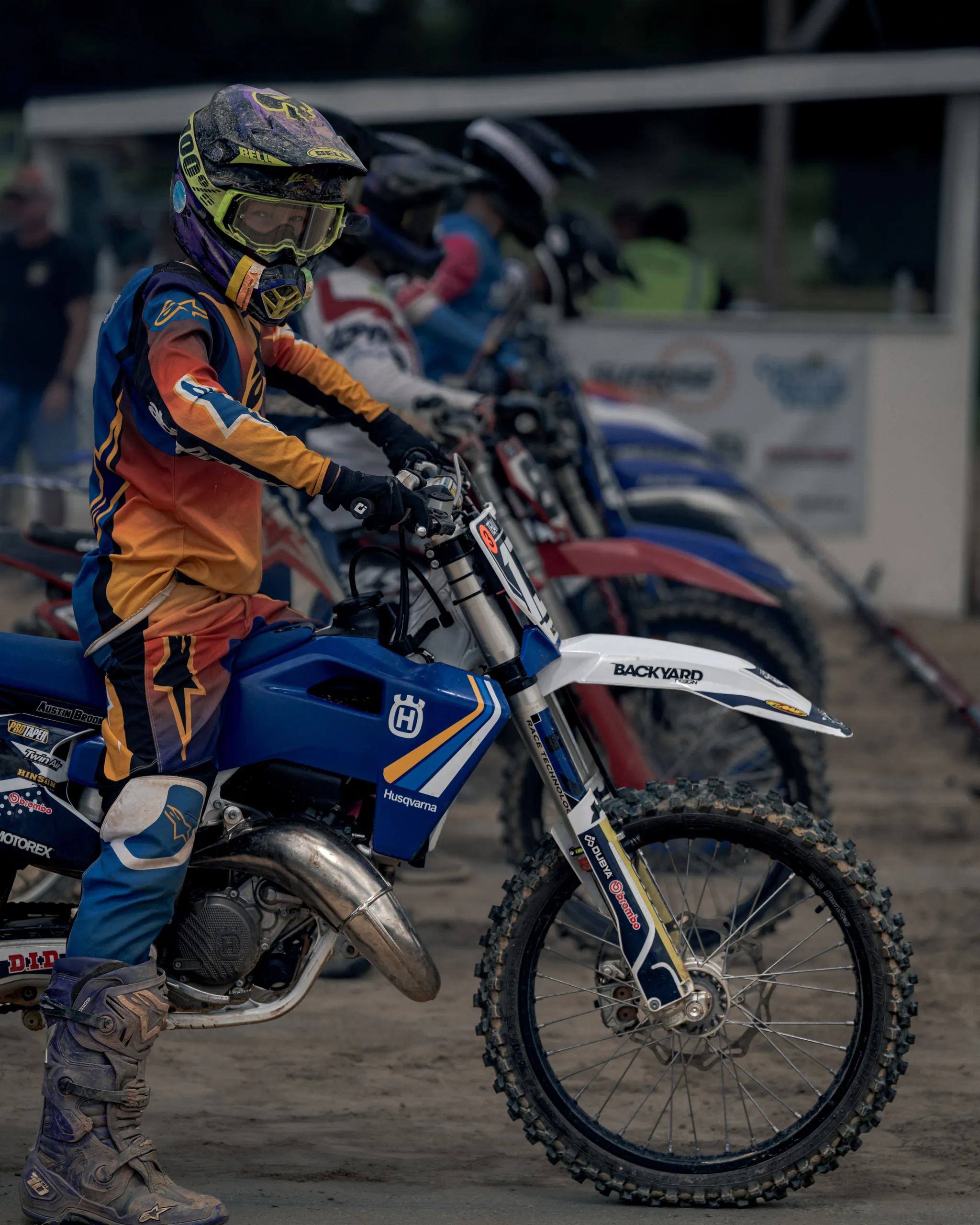 A young rider in orange and blue gear waits astride a Husqvarna at the start, a row of bikes lined up behind